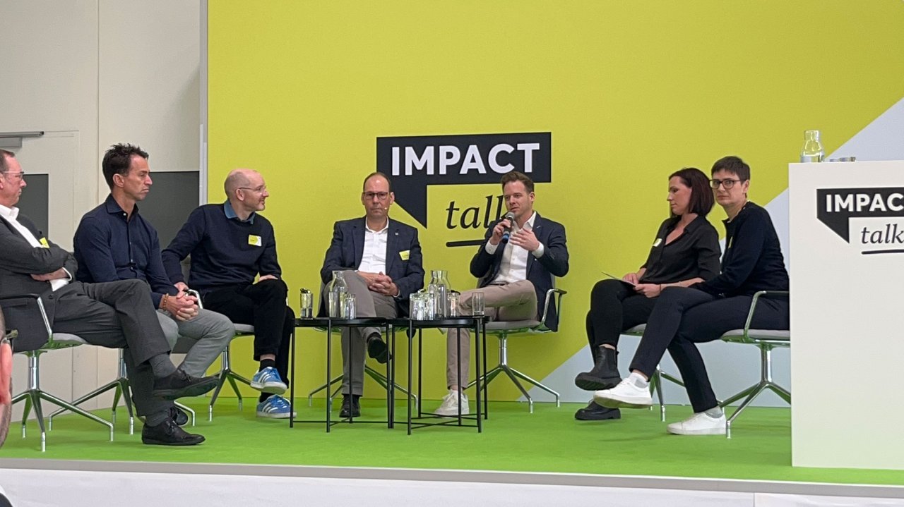 Sechs Personen sitzen auf Stühlen auf einer Bühne mit grünem Boden und gelbem Hintergrund, diskutieren bei einer Veranstaltung namens 'IMPACT talk'.