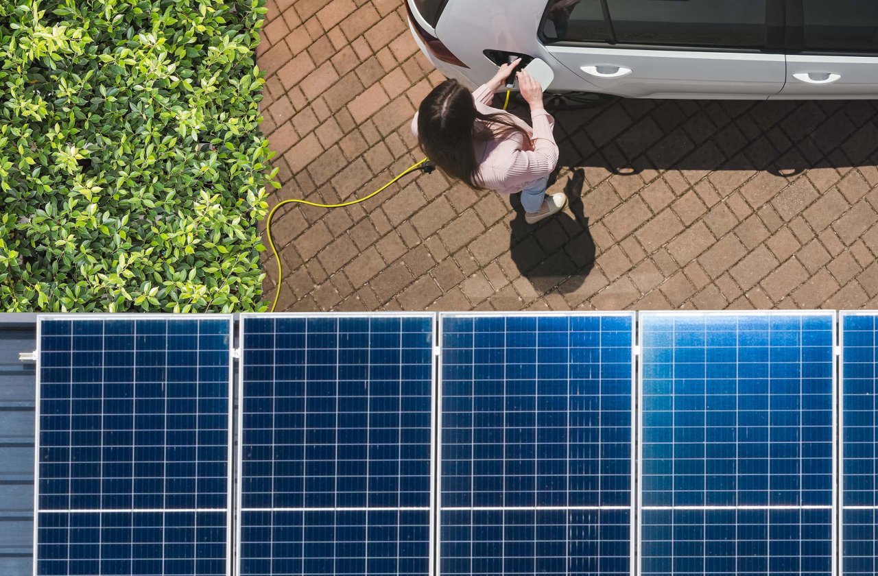 Frau lädt ein Elektroauto an einer Ladestation neben Solarpaneelen und grünen Pflanzen.