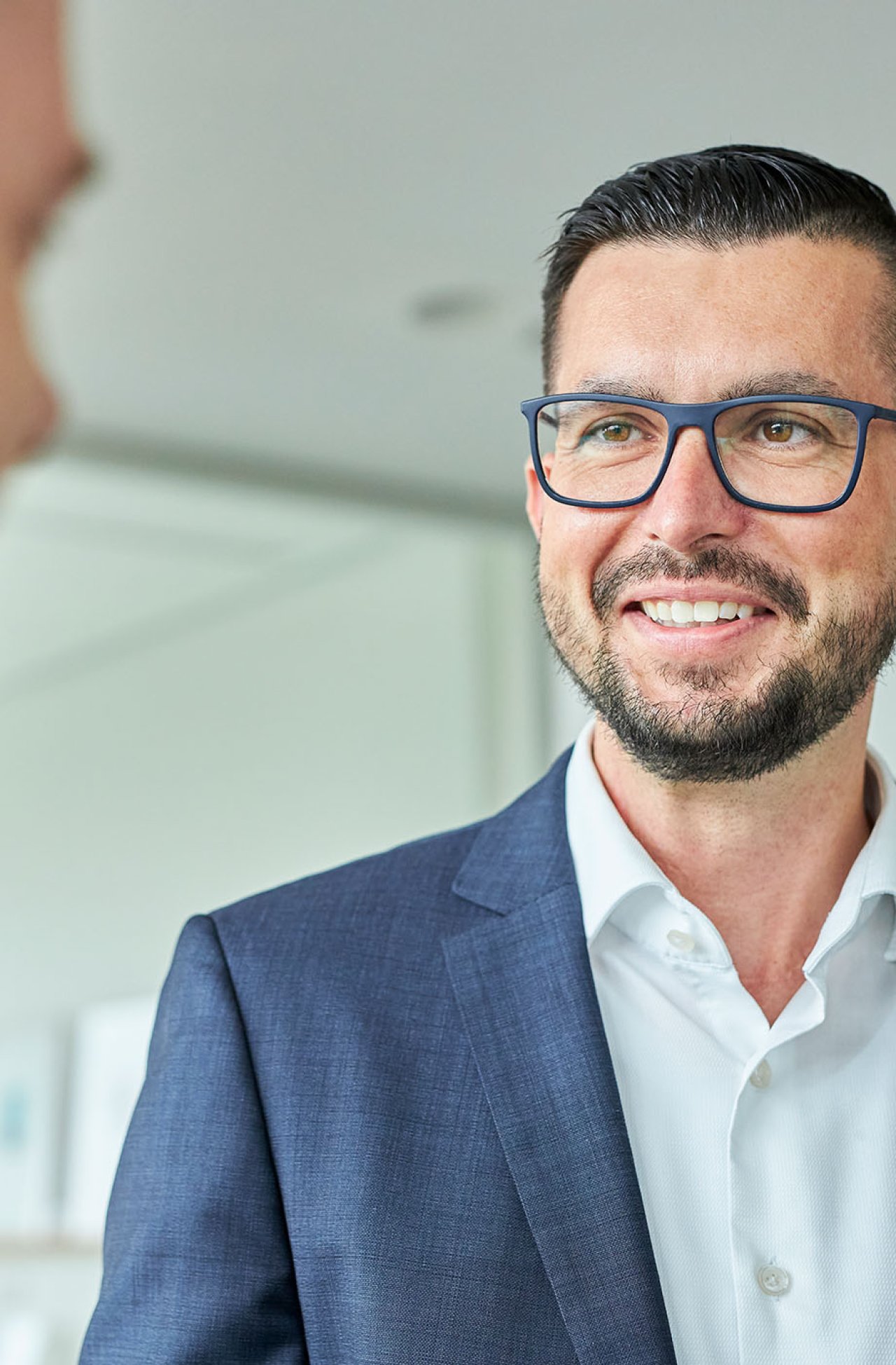 Lächelnder Mann mit Brille und Bart trägt weißen Hemd und blauen Anzug, im Gespräch in modernem Büro.