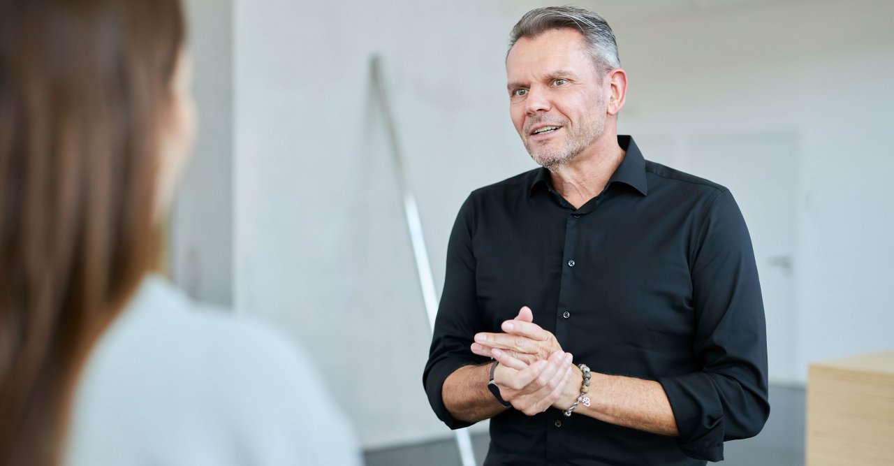 Mann im schwarzen Hemd spricht und gestikuliert mit einer Frau im Vordergrund.