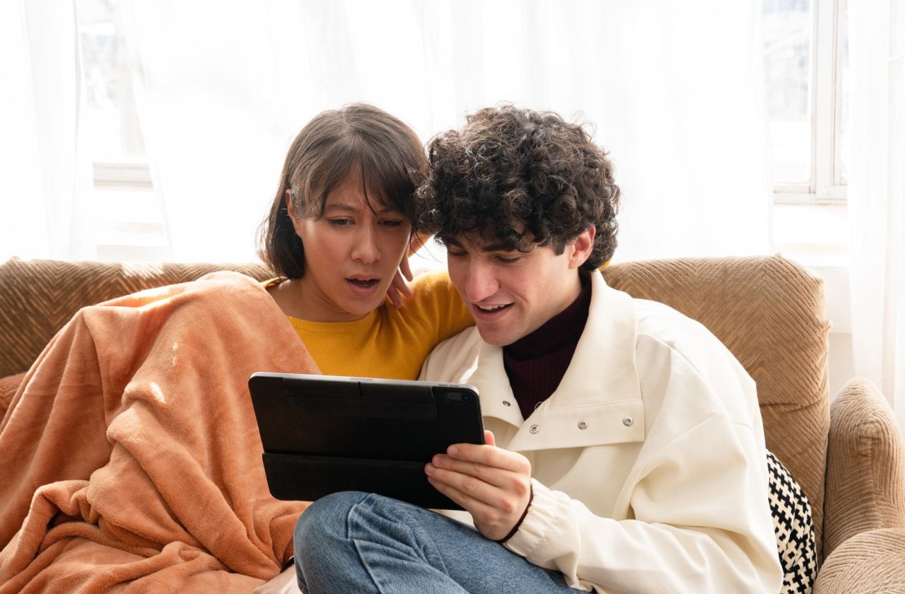 Ein dunkelhaariges Paar sitzt auf einem Sofa und schaut staunend auf ein Tablet.