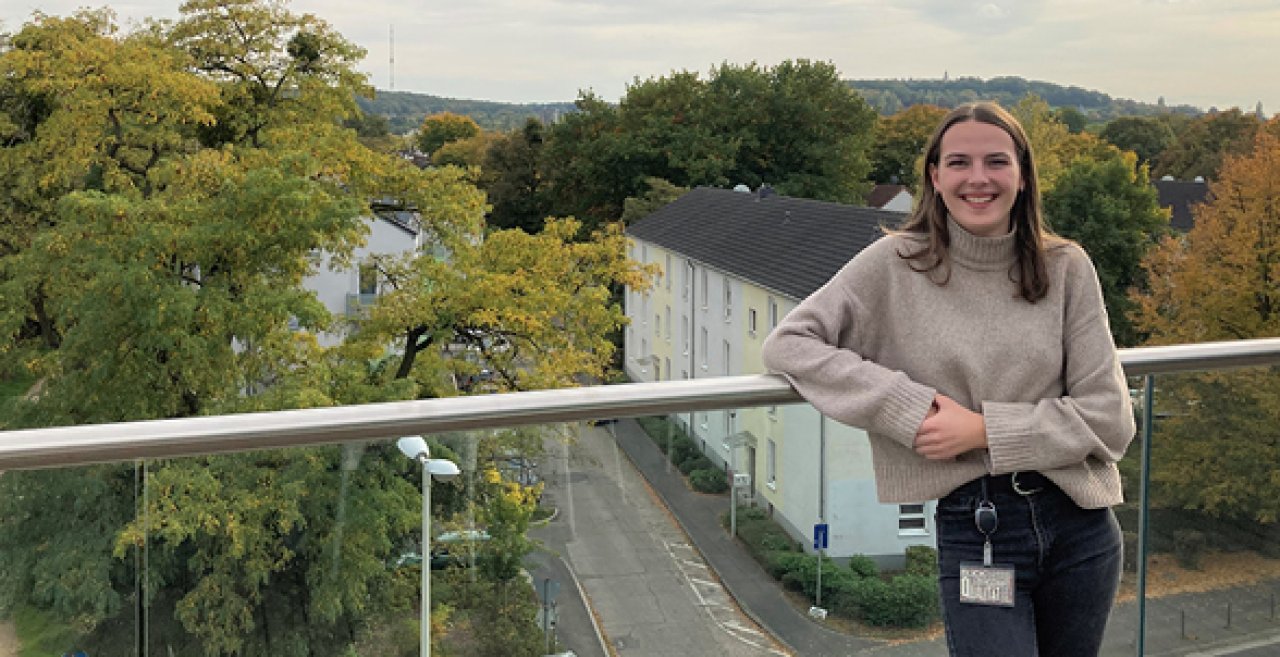 Junge Frau mit braunen Haaren und grauem Pullover lehnt lächelnd an einem Glasgeländer auf einem Balkon mit Blick auf eine Straße und herbstliche Bäume.